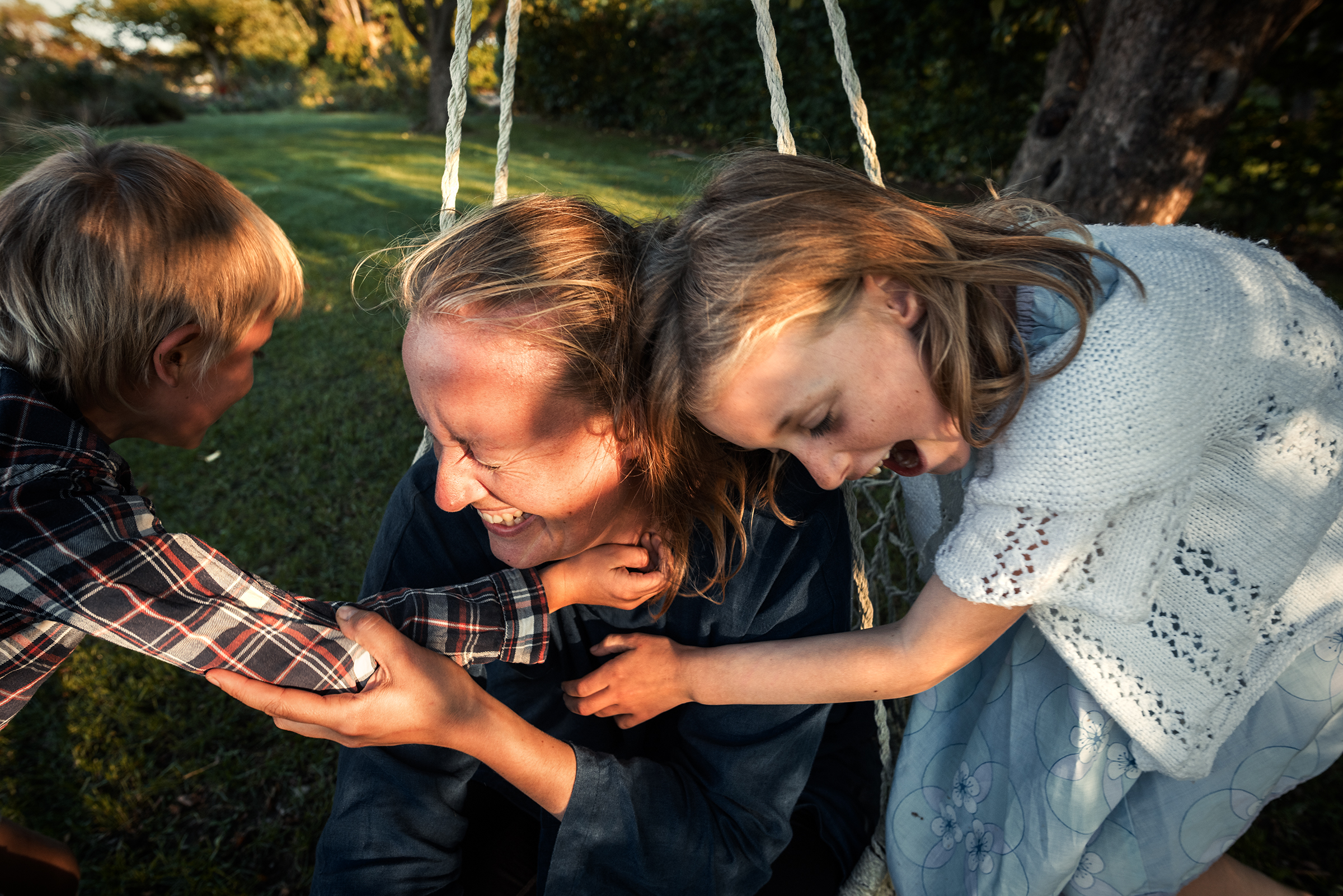 This joyful documentary family photograph captures a woman laughing in a hanging macrame chair as two children playfully embrace her, beautifully illustrating an authentic and loving family story in a sunlit garden.