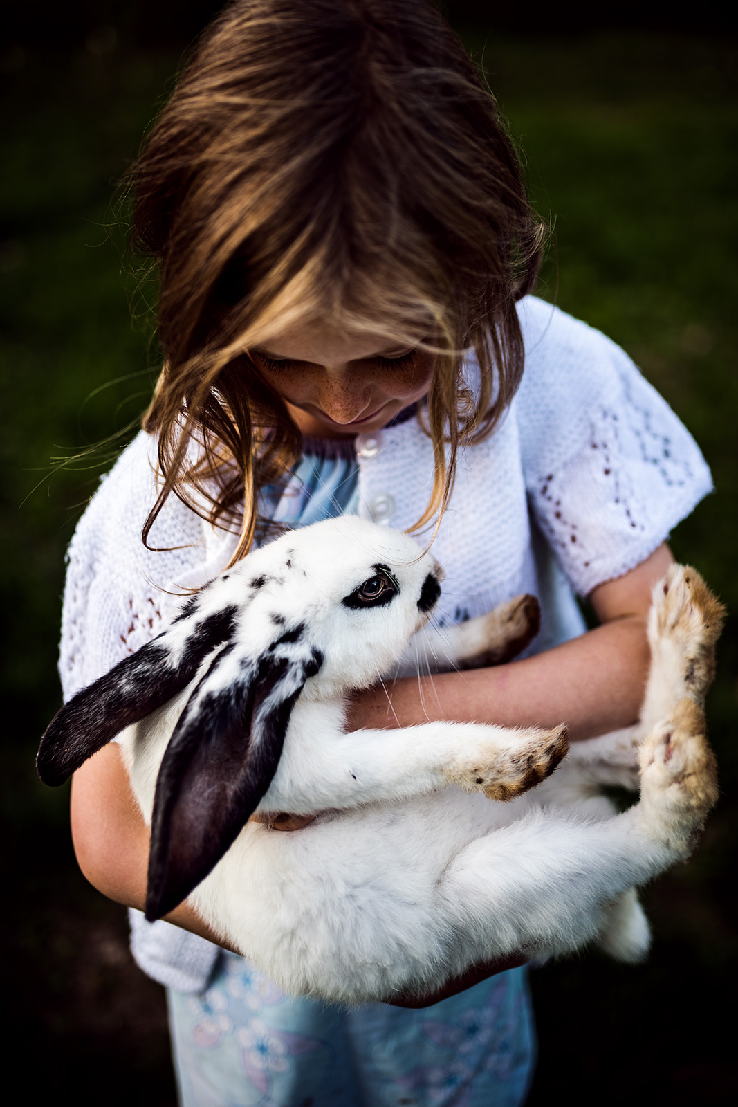 This tender documentary family photograph captures a young girl gently cradling a black and white rabbit, her gaze soft as she looks down at it, beautifully illustrating an authentic and gentle family story.
