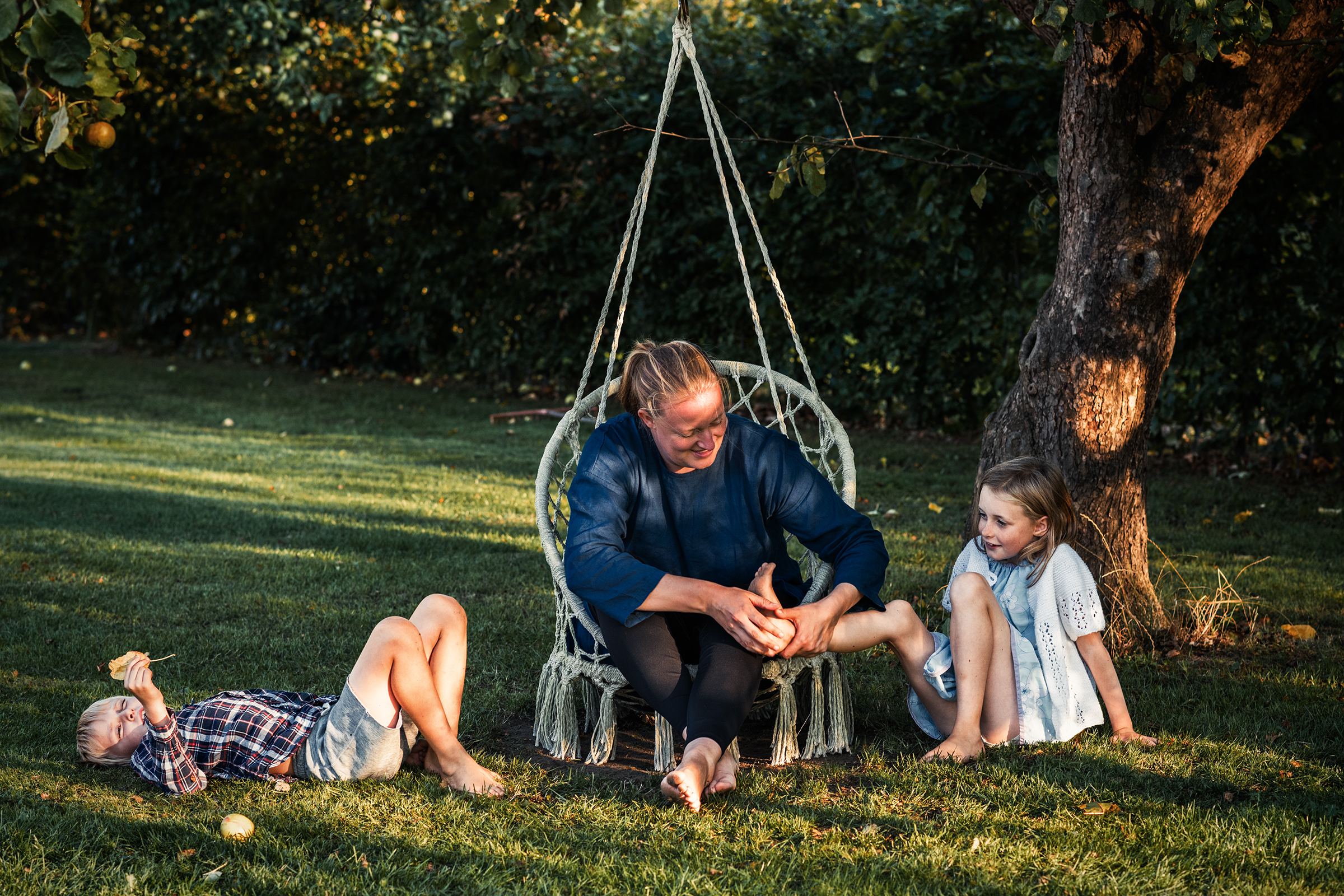 This warm documentary family photograph captures a woman in a hanging macrame chair tenderly interacting with a young girl's foot as she sits on the sun-dappled grass, while another child relaxes nearby with an apple, illustrating an authentic and playful family story.