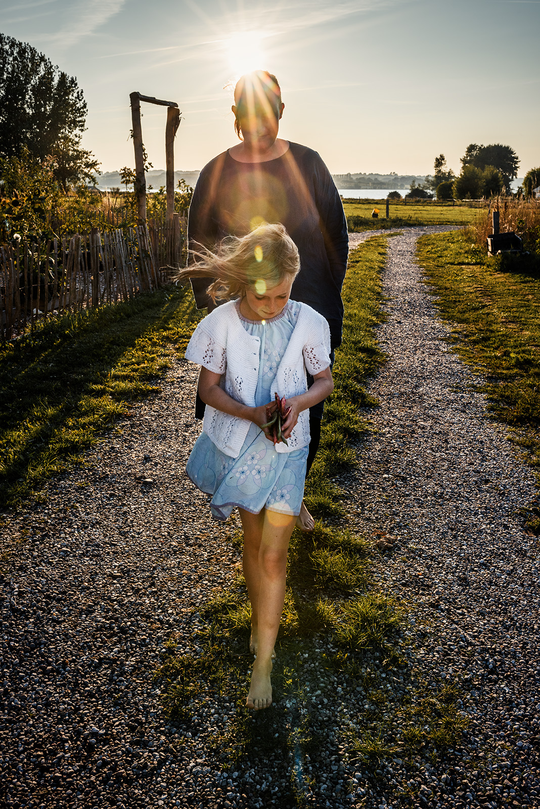 This evocative documentary family photograph captures a young girl walking barefoot on a gravel path, her hair blowing in the wind as an adult follows behind silhouetted by the bright sun, beautifully illustrating an authentic and sun-drenched family story.