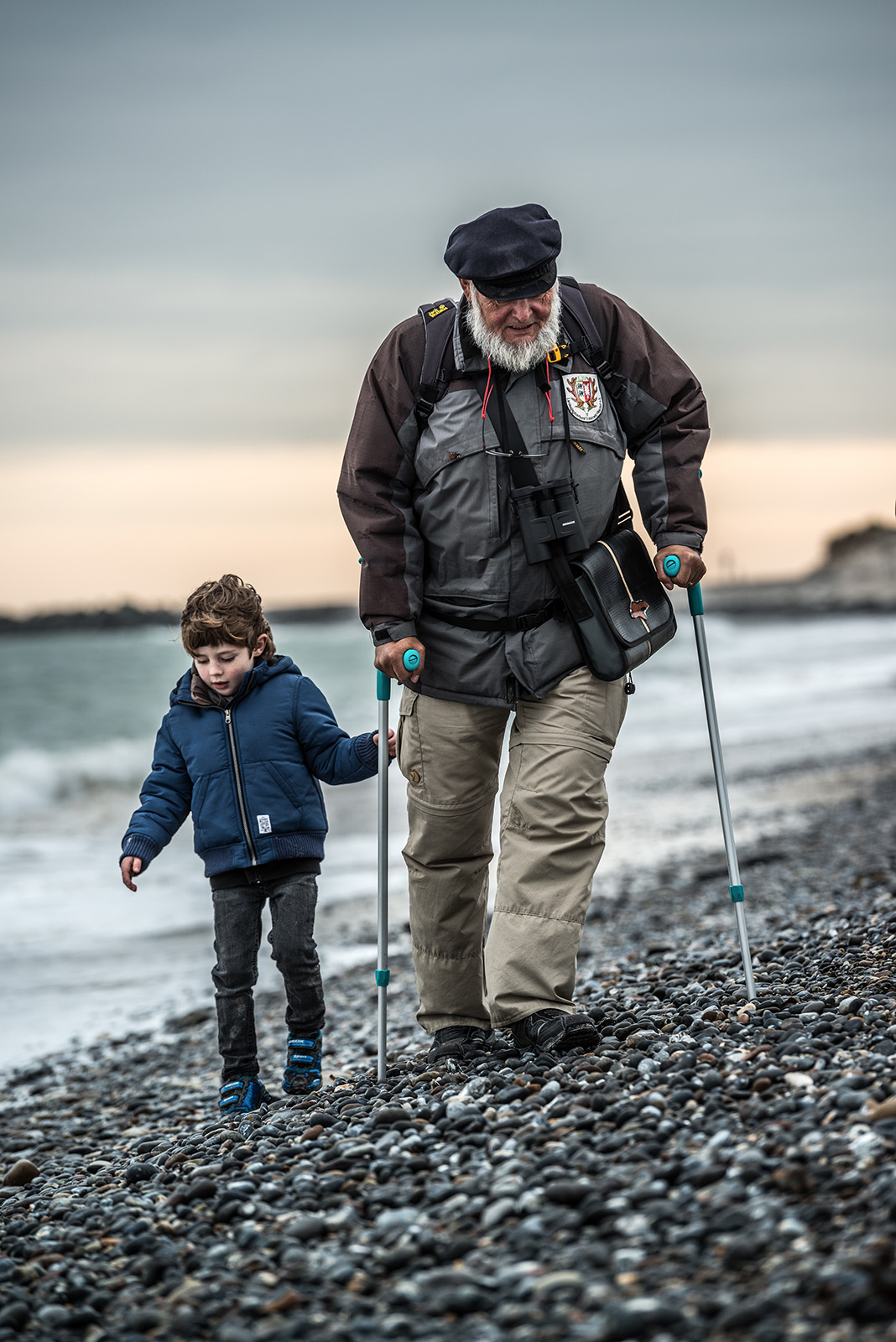 This poignant documentary family photograph captures an elderly man with crutches and a young child carefully walking hand-in-hand on a pebbly beach, beautifully illustrating an authentic intergenerational family story.