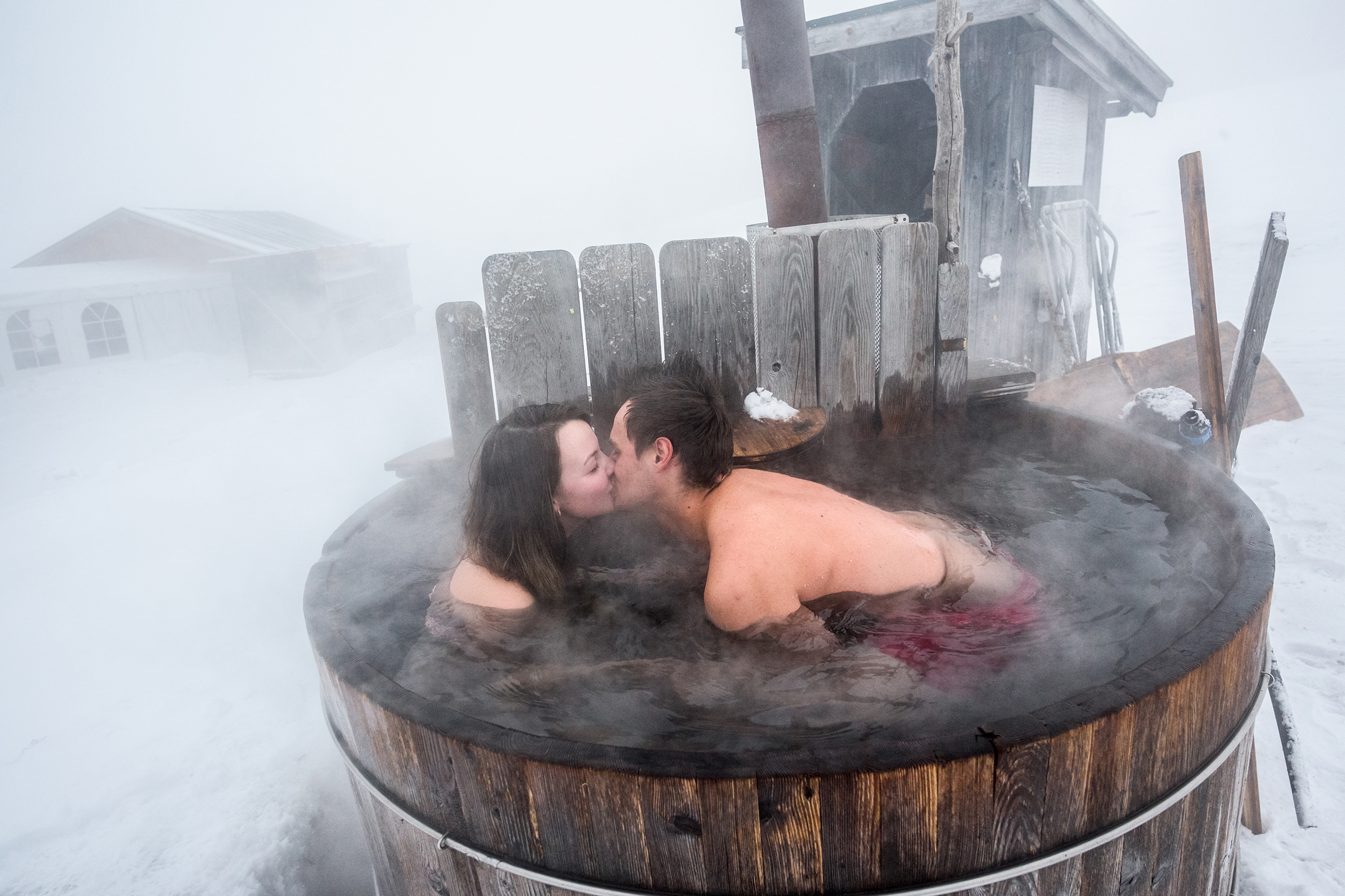 This intimate documentary photograph captures a couple kissing in a steaming outdoor wooden hot tub amidst a snowy, foggy landscape, beautifully illustrating an authentic and romantic family story.