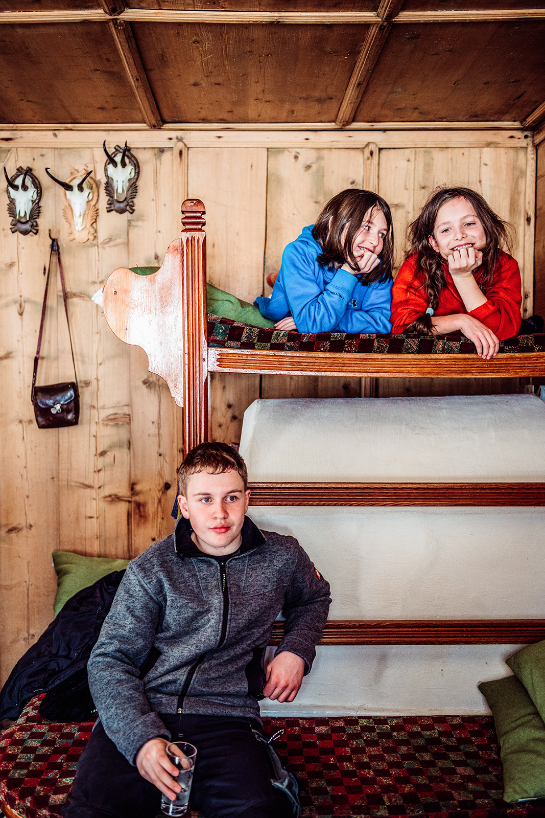 This playful documentary family photograph shows two children peeking and smiling from the top bunk of a wooden bunk bed while another child sits seriously on the bottom bunk, capturing an authentic and relatable family story.