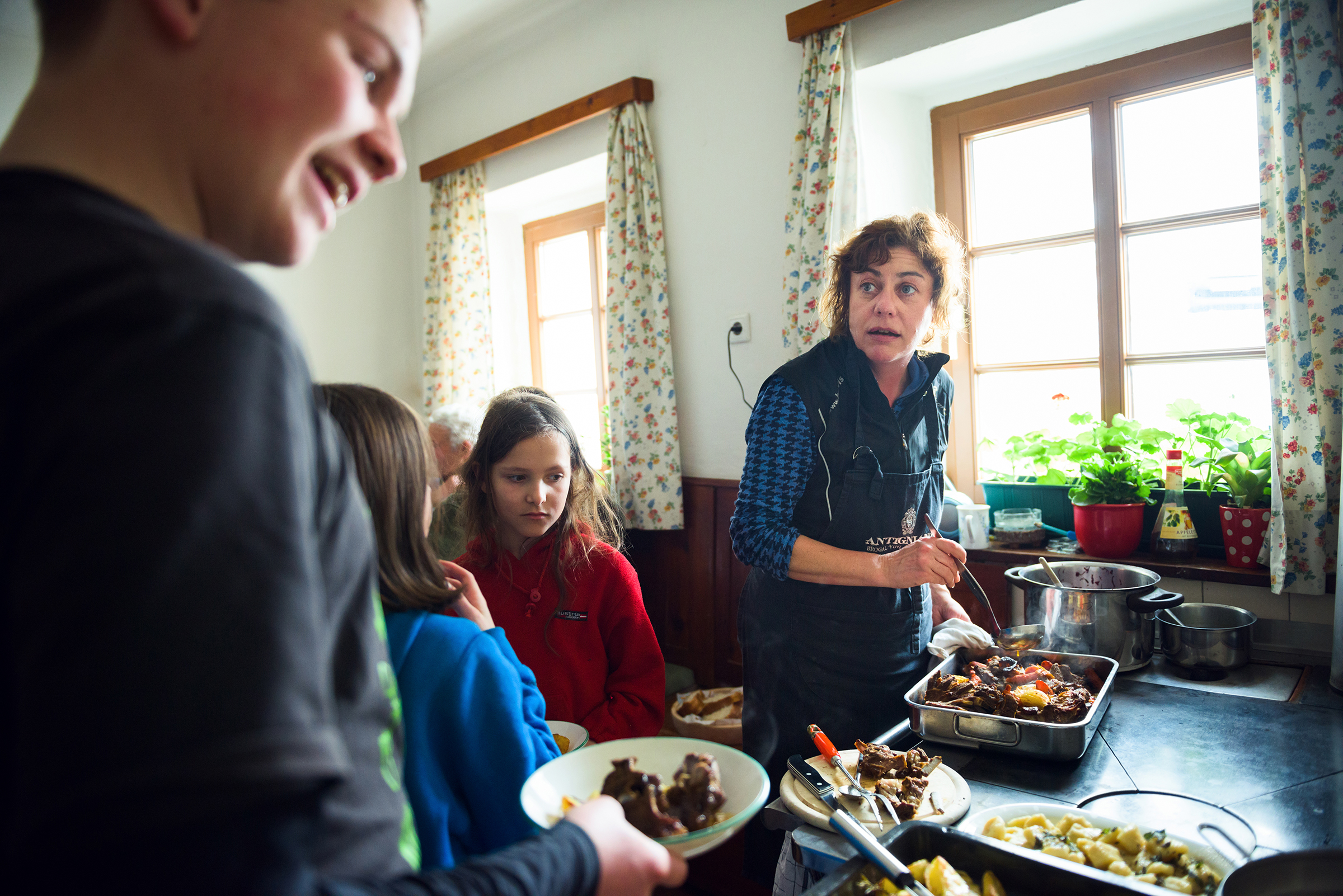 This lively documentary family photograph captures a woman in an apron serving a hearty meal in a sunlit kitchen while children wait, perfectly illustrating an authentic family gathering story.