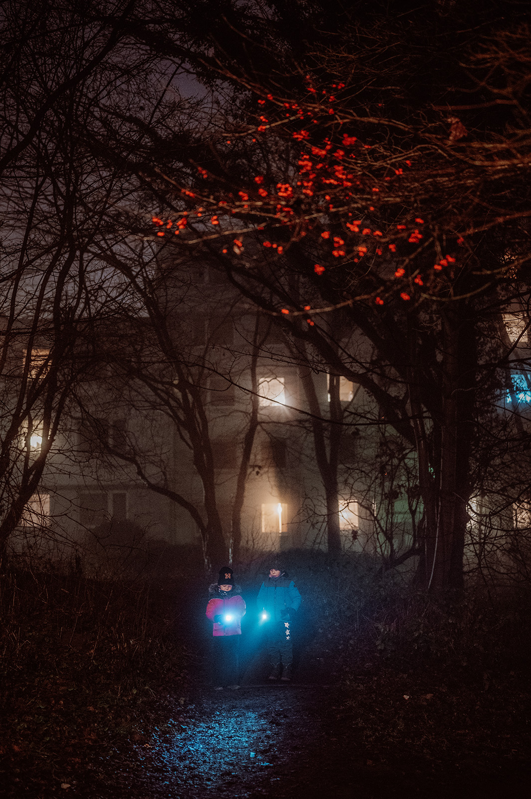 This atmospheric documentary family photograph captures two children with bright flashlights on a nocturnal adventure through a misty, wooded area, with a warmly lit building and red berries framing their journey, beautifully illustrating an authentic family story.