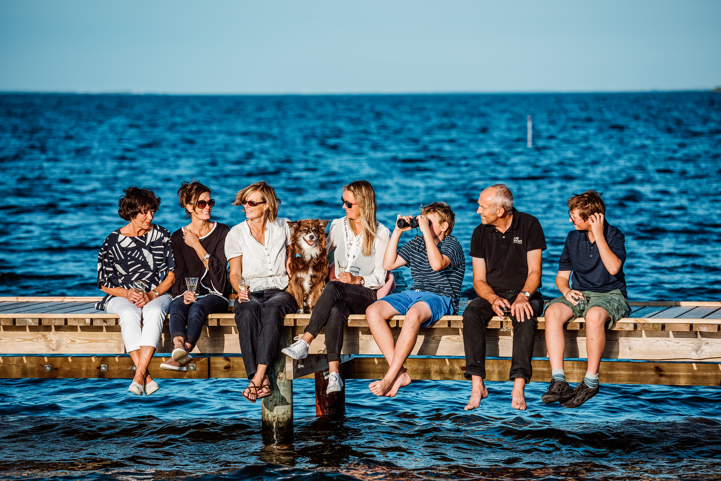 This vibrant documentary family photograph captures a multi-generational family and their dog enjoying a sunny day on a wooden pier over deep blue water, sharing drinks and conversation as one boy looks through binoculars, perfectly illustrating a cherished, authentic family story.
