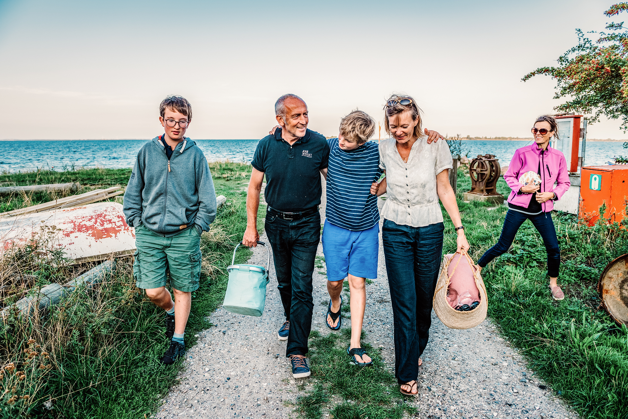 This vibrant documentary family photograph captures a multi-generational family walking happily together along a coastal path, carrying a bucket and basket, beautifully illustrating an authentic shared story.