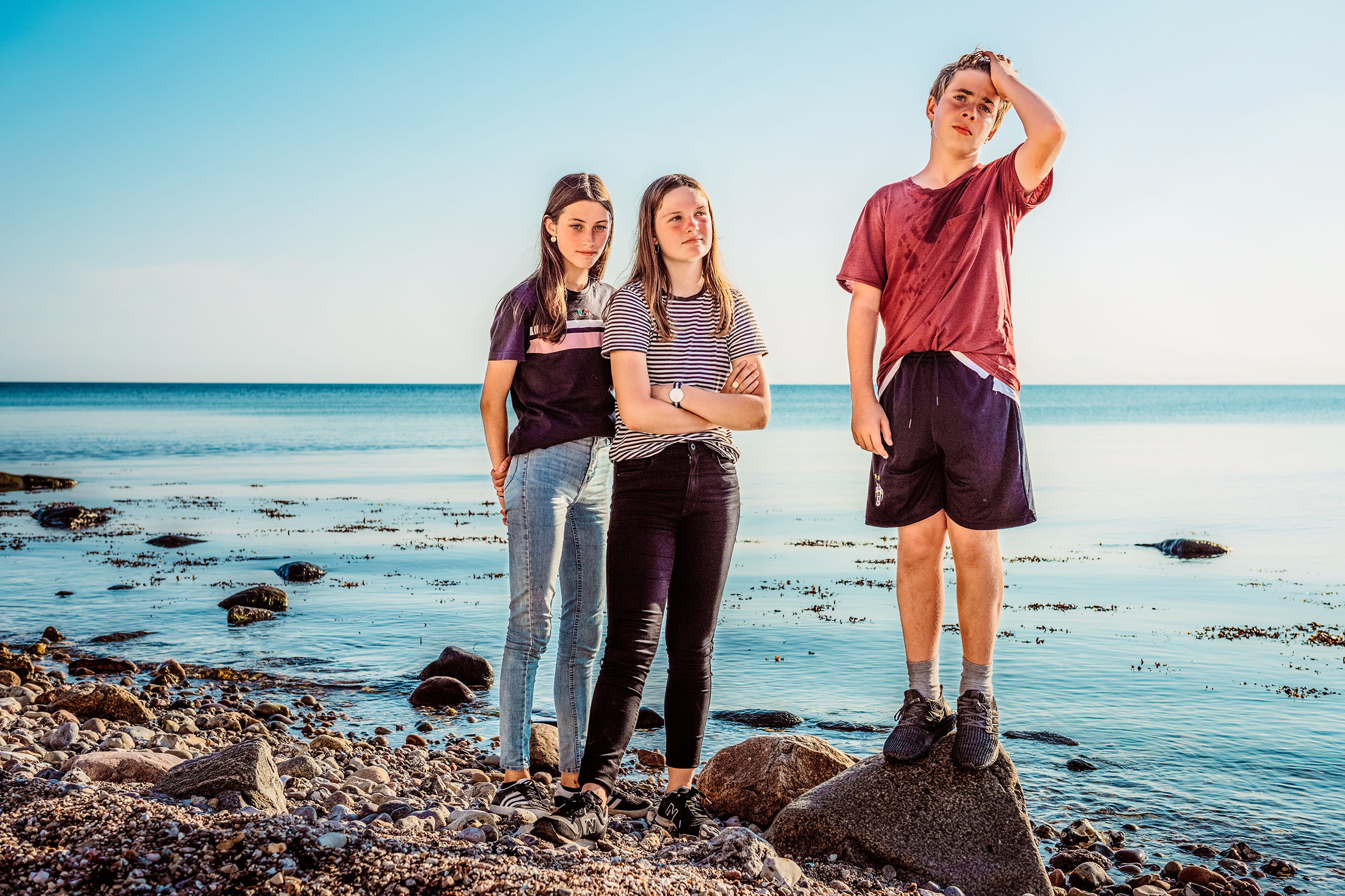 This vibrant documentary family photograph showcases three young people—two girls standing together and a boy on a rock—against a serene sea and bright sky, capturing an authentic family story.