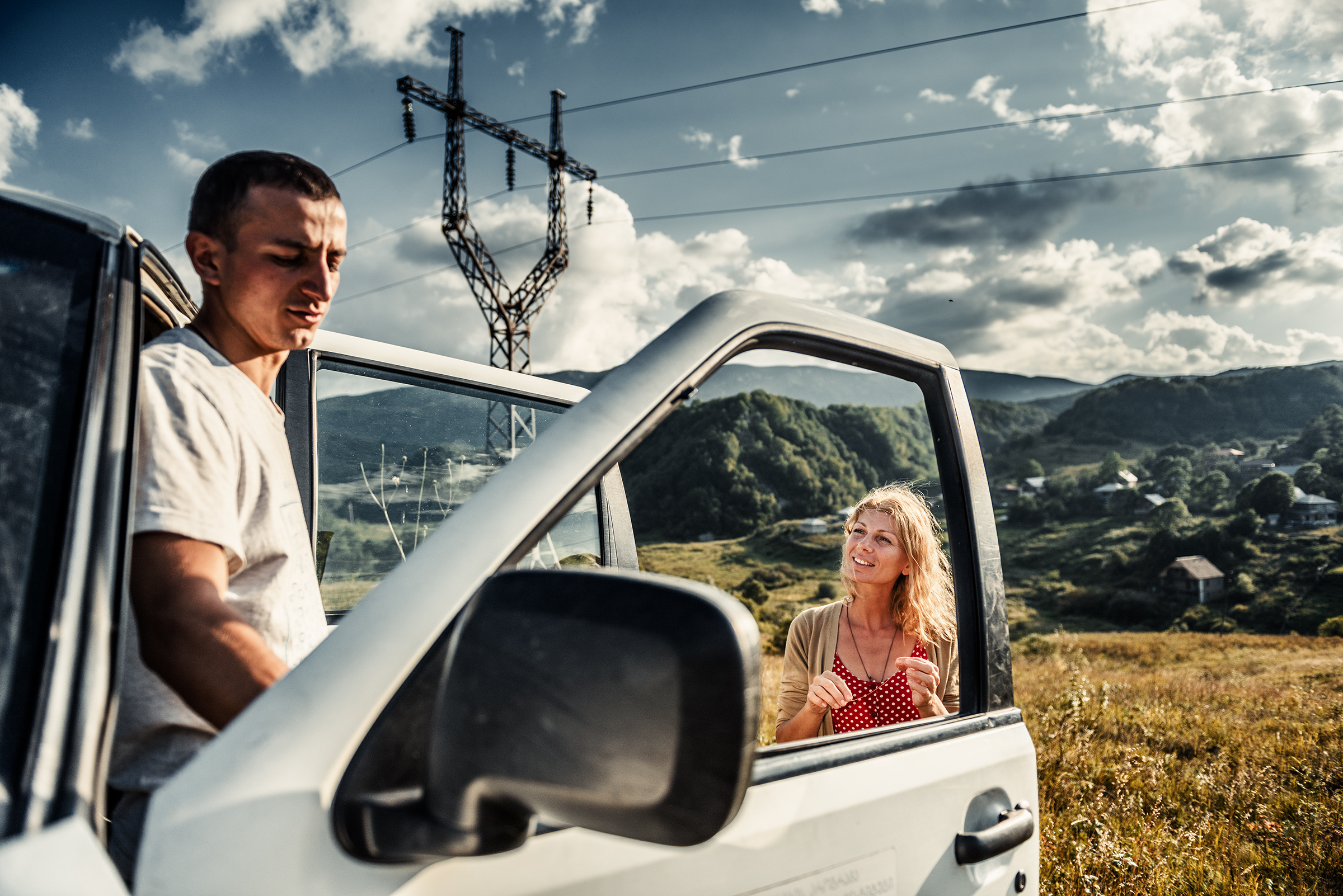 This atmospheric documentary family photograph shows a man by the open driver's door and a woman standing outside, set against a dramatic rural landscape with an electricity pylon, illustrating an authentic travel story captured through storytelling photography.