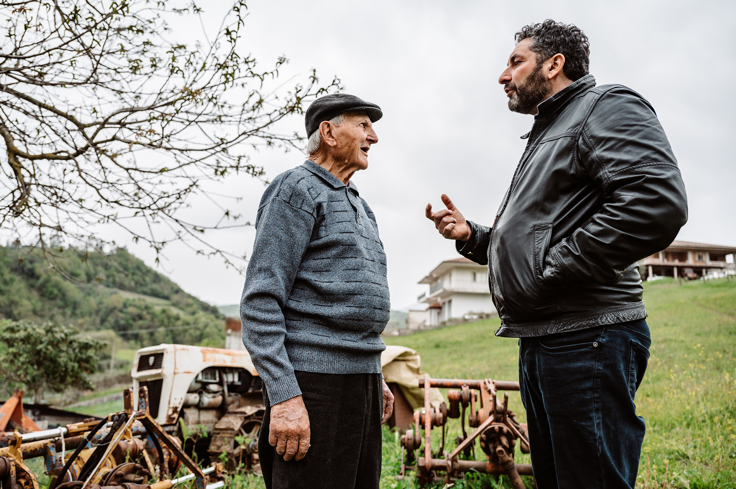 This compelling documentary family photograph captures an elderly man in a cap and a younger man in a leather jacket engaged in earnest conversation amidst rustic farm equipment, perfectly illustrating the authentic storytelling of intergenerational family connections.