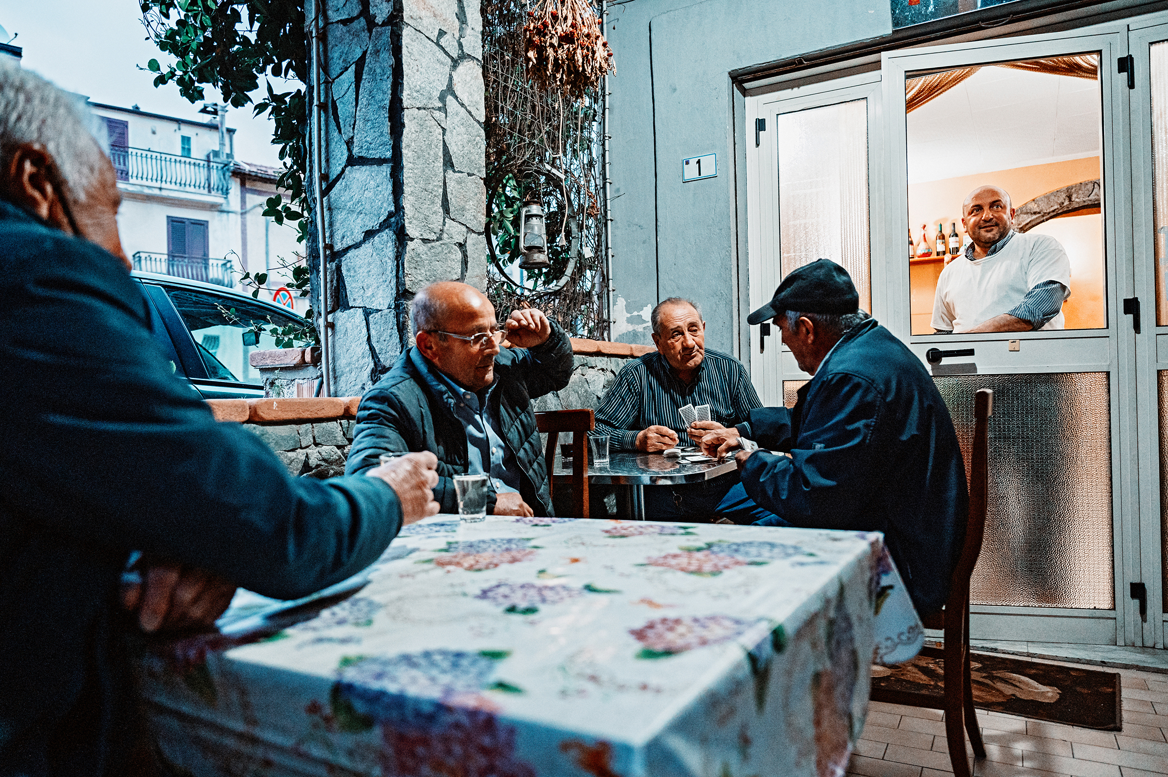 This candid documentary photograph captures four older men absorbed in a card game and conversation at an outdoor cafe table, with another man observing from a window, perfectly illustrating the authentic storytelling of documentary and family photography.
