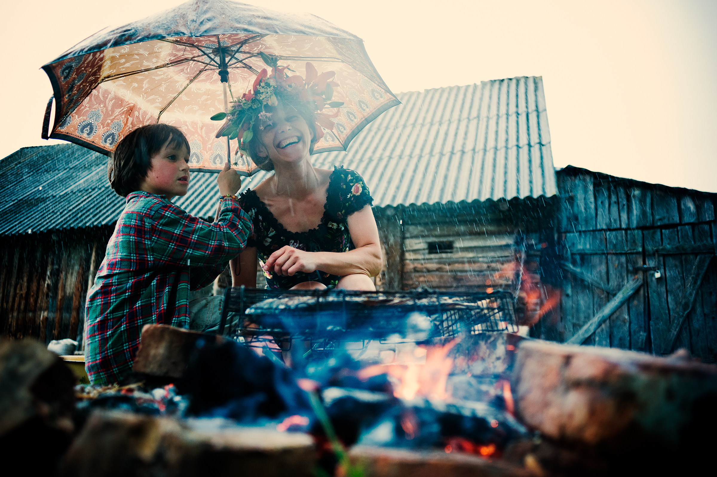 This joyful documentary family photograph captures a woman wearing a flower crown and a child sharing an umbrella by a glowing campfire in the rain, beautifully illustrating an authentic and memorable family adventure.