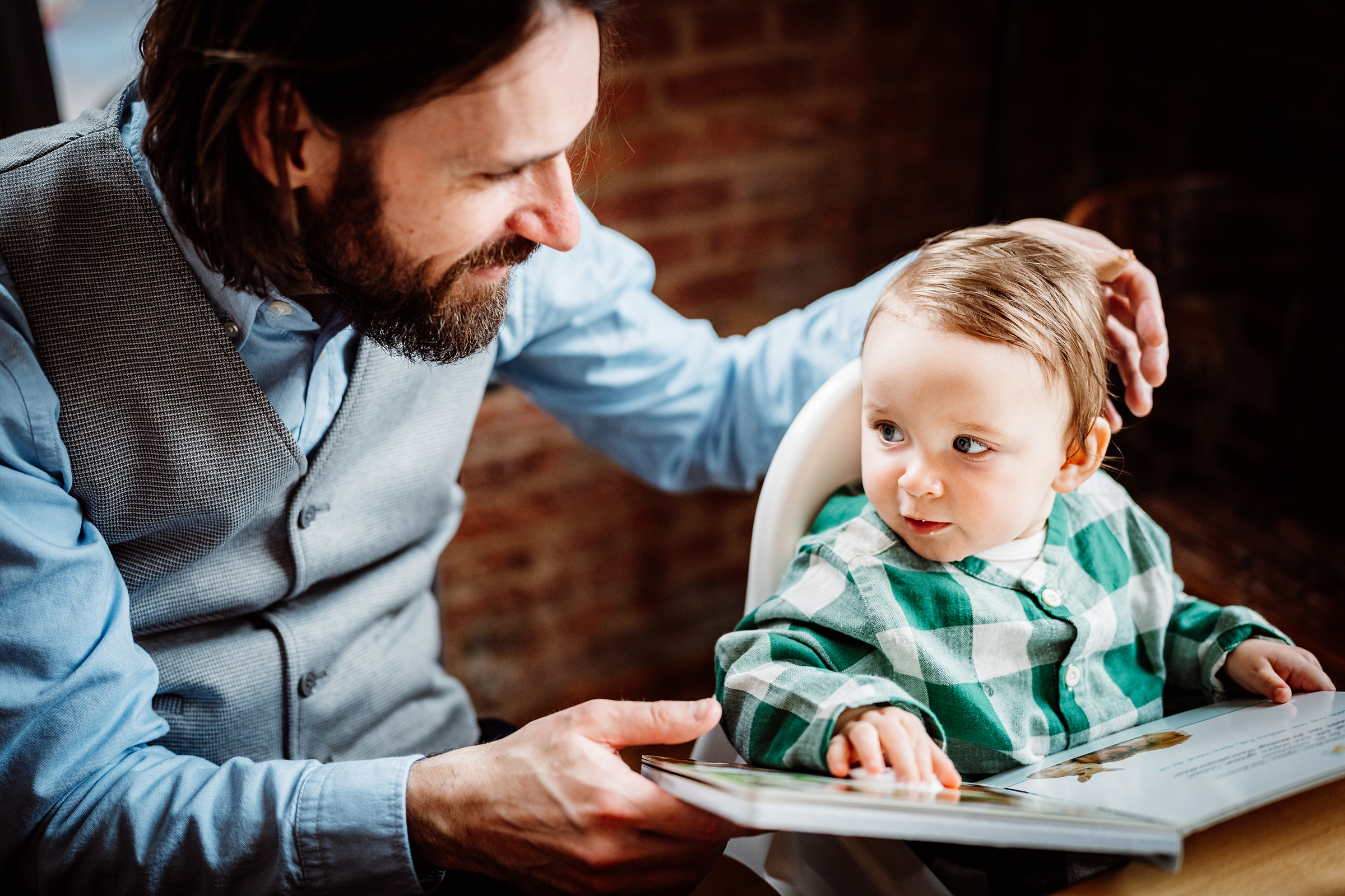 This heartwarming documentary family photograph captures a father tenderly engaging with his young child in a highchair over a picture book, beautifully illustrating an authentic family story.