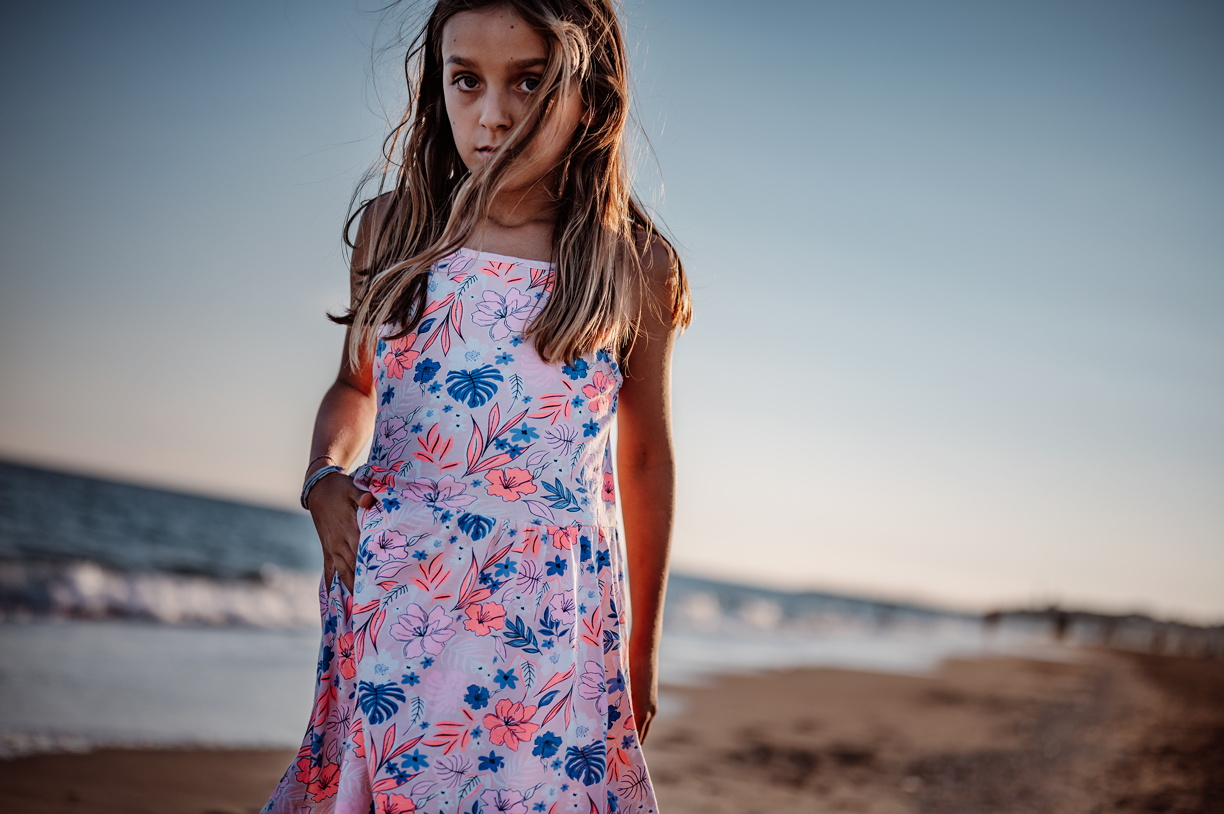 This compelling documentary family photograph captures a young girl in a floral dress on the beach at golden hour, her direct gaze reflecting an authentic childhood story.