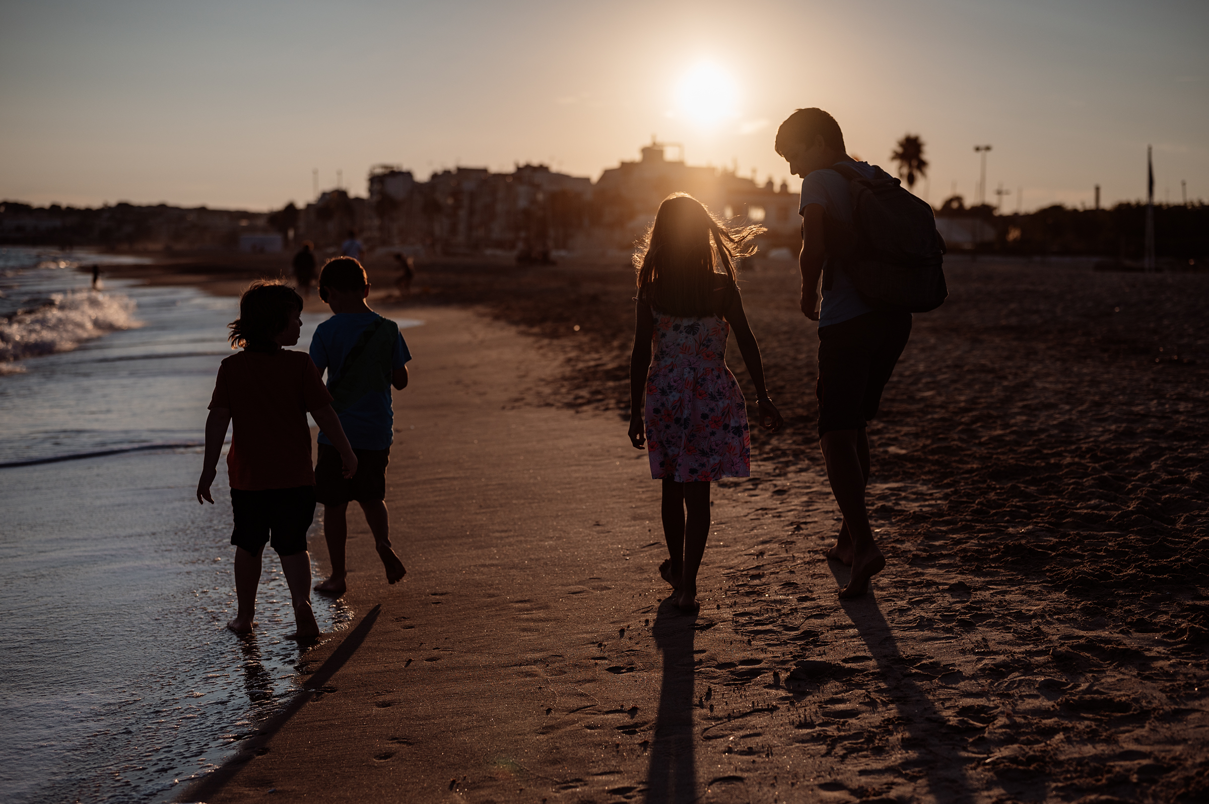 A documentary family photograph shows a father and three children silhouetted as they walk along the beach at sunset, capturing an authentic and cherished family moment.