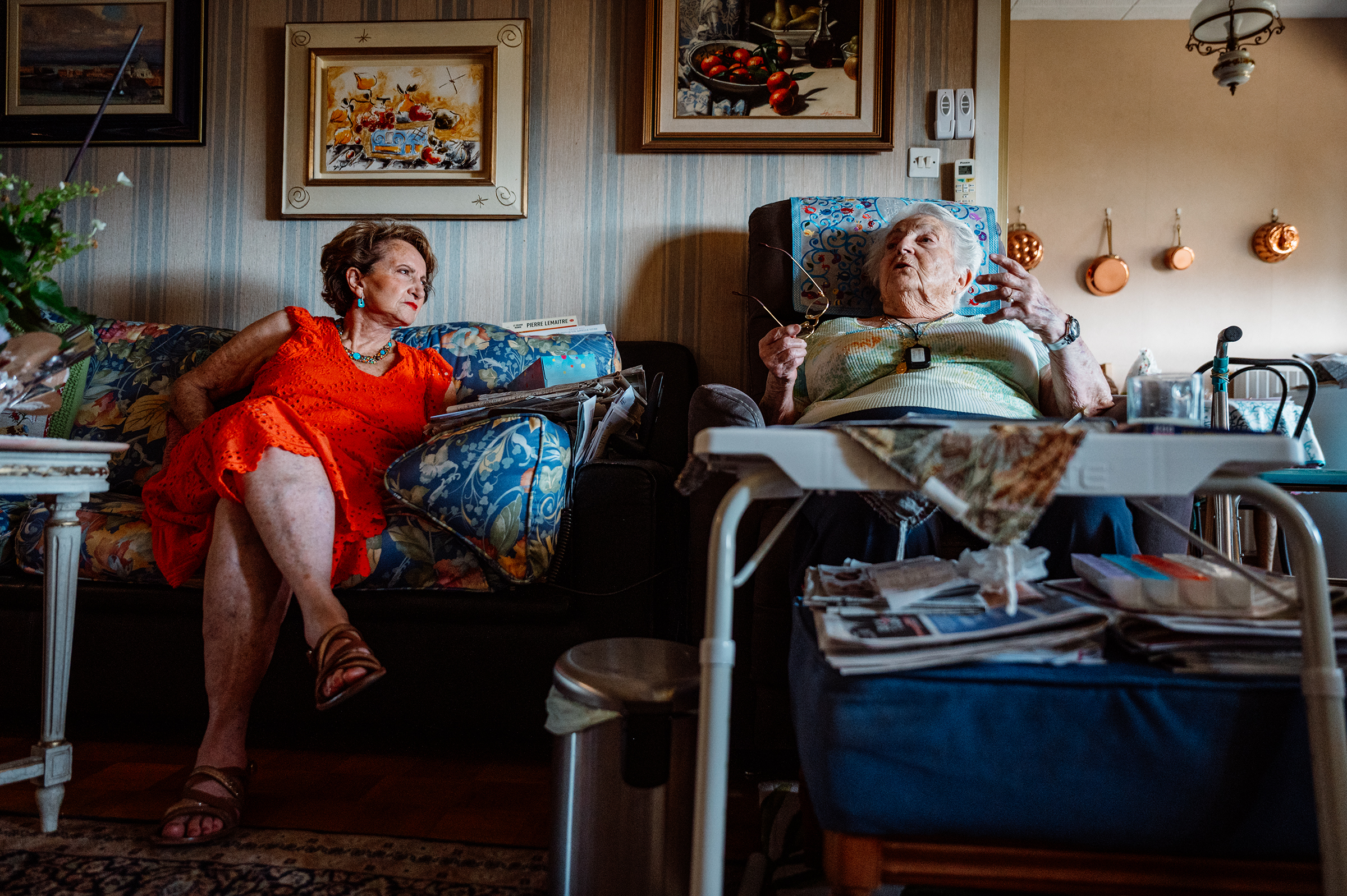 A candid documentary family photograph captures an authentic interaction between an older woman in her recliner and a younger woman on the sofa in a characterful living room, showcasing the storytelling power of family photography