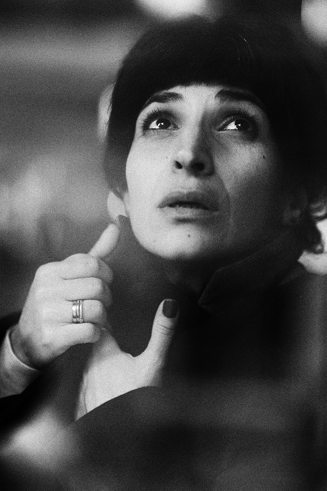 Black-and-white documentary-style portrait of a woman gazing upward with a thoughtful expression, her hands near her face, conveying introspection and emotional depth.